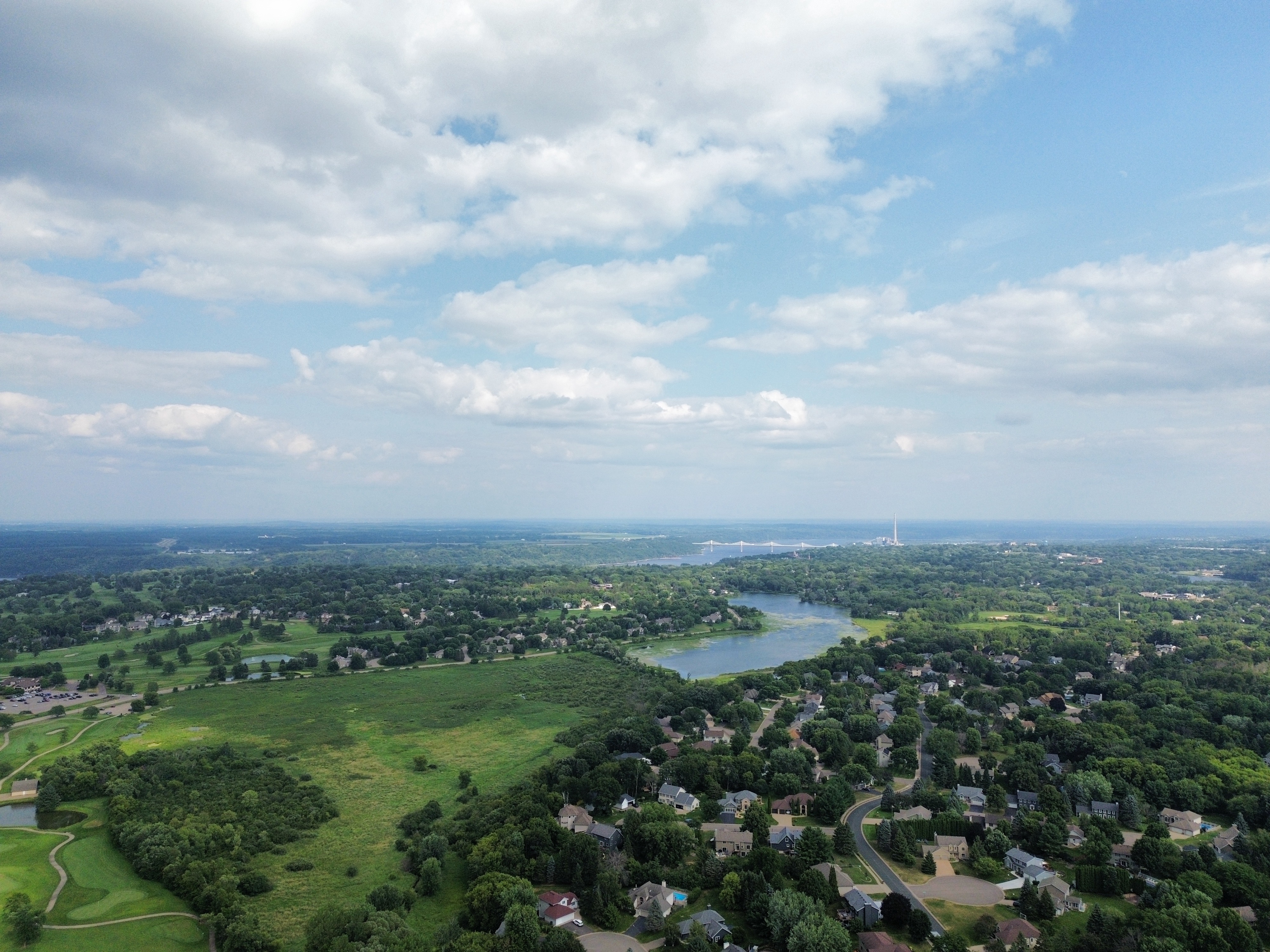 Wide aerial view over homes, golf fairways, and a weedy lake stretching toward a distant bridge